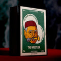 A matchbox-style product box standing upright on a red surface against a dark smoky background. The front label reads 'The Wrestler' and shows a stylized illustrated face with a red hat and gold/yellow details on a green panel.