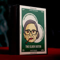 A  'The Elder Sister' matchbox , standing upright on a red surface against a dark, smoky background. The packaging features a green and white design with a stylized illustration of an elderly woman wearing glasses and smoking a cigar.